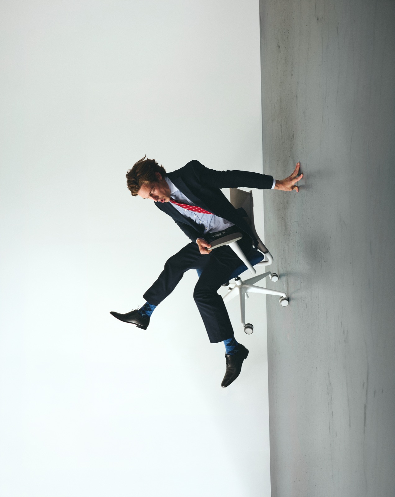 Isaac in a suit and tie riding an office chair sideways off a wall in the grey studio