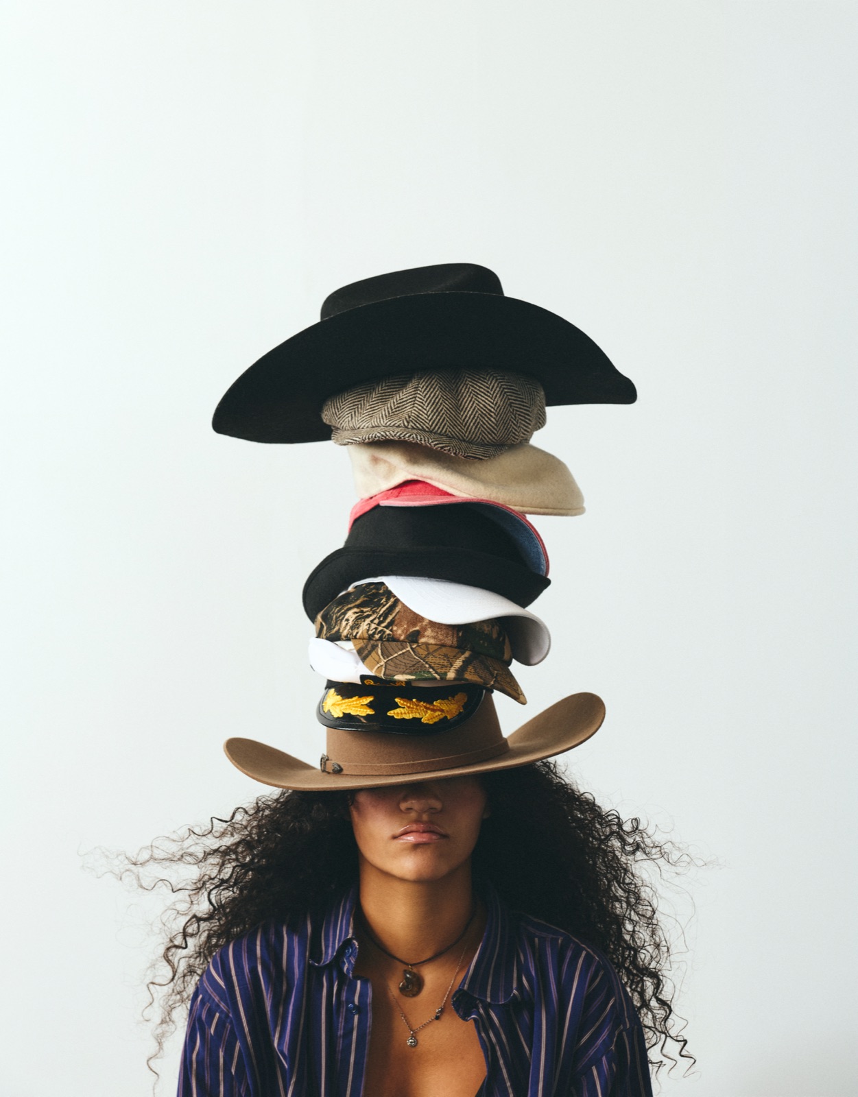 Malia under a towering stack of hats, cowboy hat on top, the brim of the last one pulled down over her eyes
