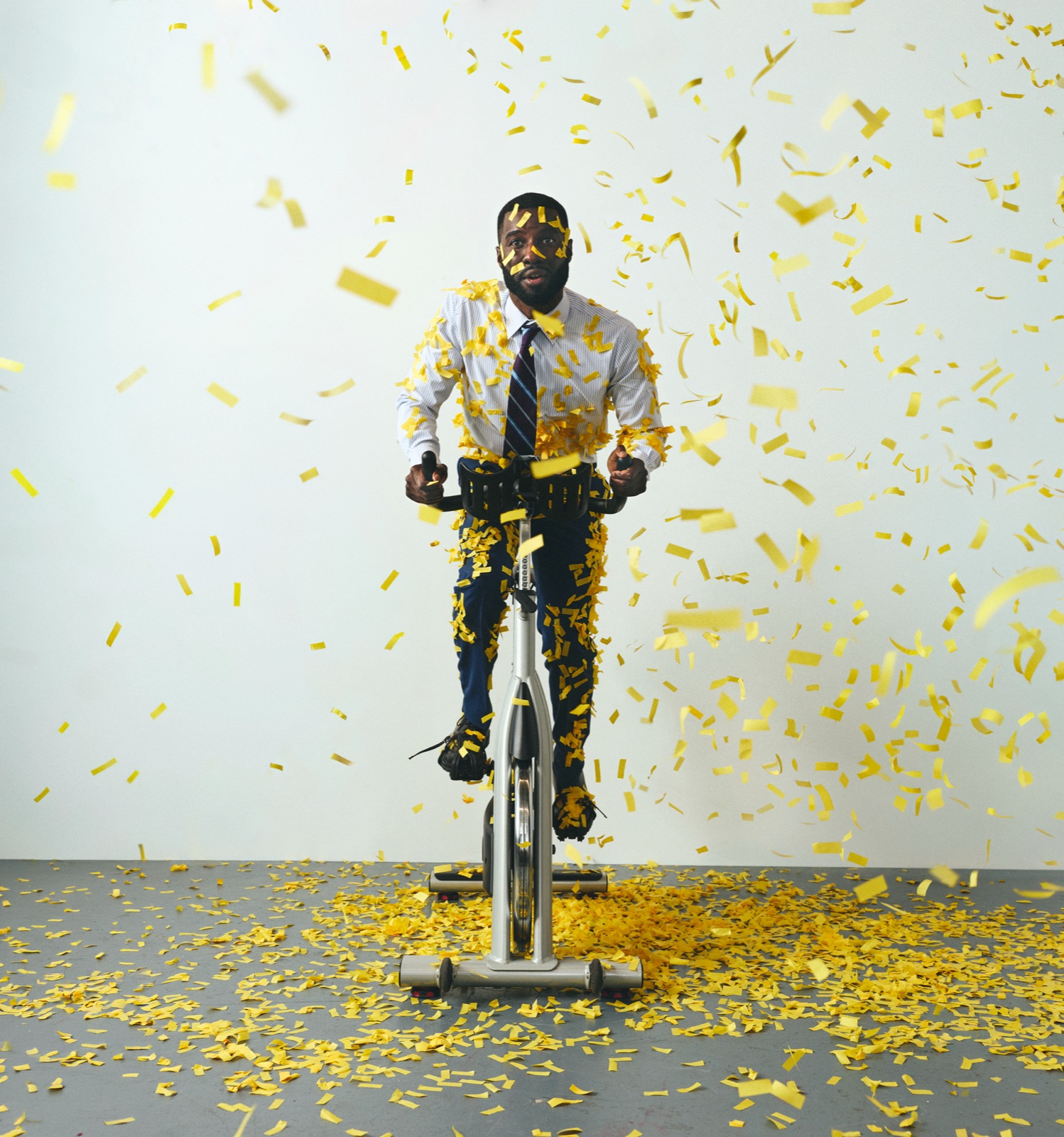 Marcus in shirt and tie on a spin bike, a storm of gold confetti falling around him in the grey studio