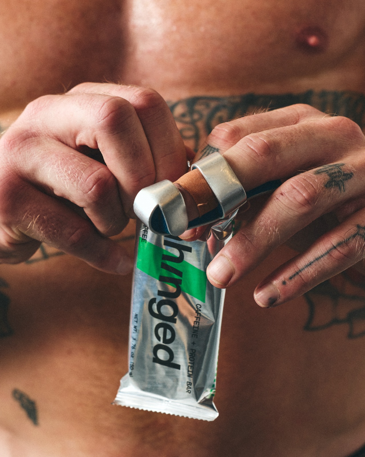 Austin, shirtless and tattooed, tearing open a foil Unhinged bar wrapper