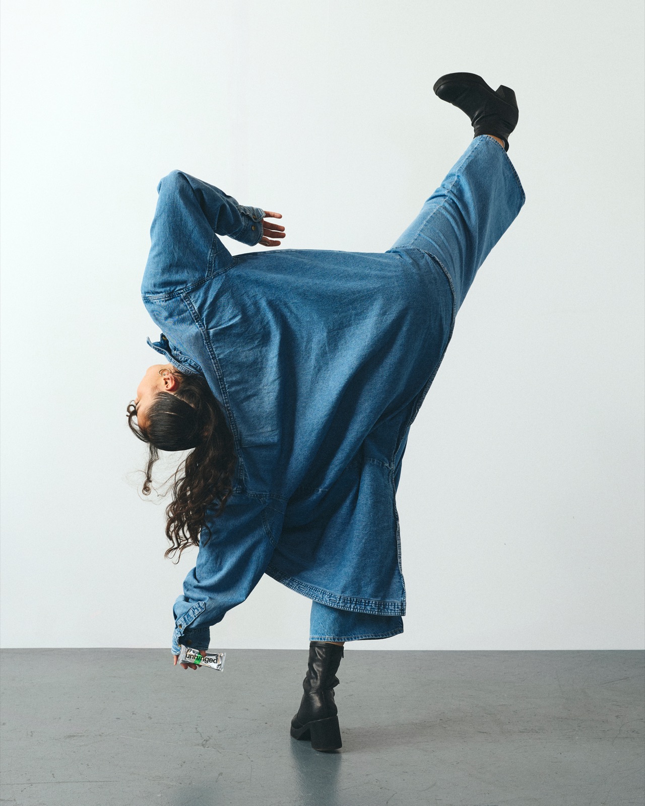 Mikayla in head-to-toe denim mid-kick, one leg straight up, an Unhinged bar in hand